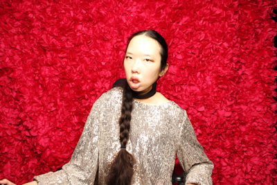 Aubrie looks at the camera from within a photobooth. Her hair is in a braid, and she wears a glittering dress, a black choker, and gold ear jewelry.
